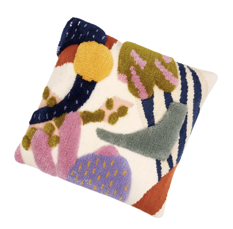 A top-down, slightly angled shot of a square throw pillow with a highly textured, abstract design on an off-white background. The design features various shapes, colors, and textures, many appearing to be tufted or embroidered with raised surfaces. Starting from the top left and moving clockwise: In the top left corner, there's a dark blue, irregular, almost boomerang-like shape. To its right, a bright yellow, roughly circular shape, appearing very fluffy or tufted. Below the yellow shape, a dark blue, curved, thick "C" or crescent shape. Further right, in the top right quadrant, there's an olive green, irregular blob shape with several short, lighter pink lines or dashes emanating from its top edge. To the right of that, a section of vertical dark blue and cream-colored stripes. Below the stripes, a prominent, raised, light teal or sage green, irregular blob shape that tapers to a point on the right. In the bottom right corner, a section of burnt orange or rust-colored material with a subtle diagonal pattern. Moving to the bottom center, a purple, somewhat rounded, irregular shape covered in small, lighter purple or pink dots. To the left of the purple shape, a cluster of pink, elongated, rounded shapes that appear very fluffy or tufted, resembling flower petals or thick fingers. Above the pink shapes, a dark olive green, irregular shape with several small, lighter yellow or beige dots on it. This shape appears to be overlapping the yellow circle mentioned earlier. The overall aesthetic is vibrant, playful, and tactile, reminiscent of modern art or abstract landscapes.